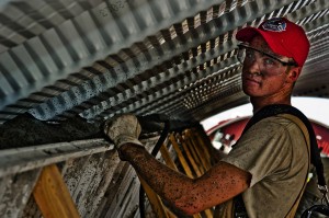 A young man working in construction. He is covered in dirt, wearing safety glass, a red cap and a safety harness.