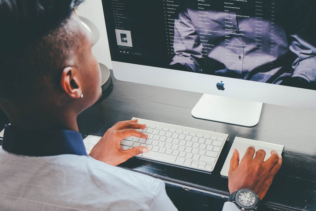 A man sitting at his desk in a home office, working on his computer. He is reflected in the monitor.