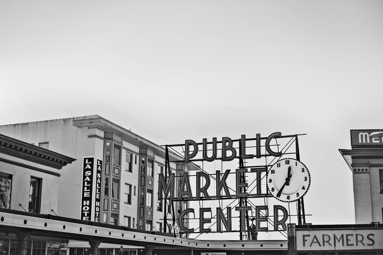 A black and white photograph of Pike Place Public Market in Seattle, with a large sign with a clock on it.