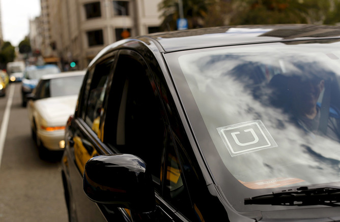 A black car on a busy street with the Uber logo in the corner of the windshield.