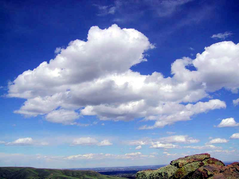 Cumulus clouds in a blue sky over hills and rock formations.