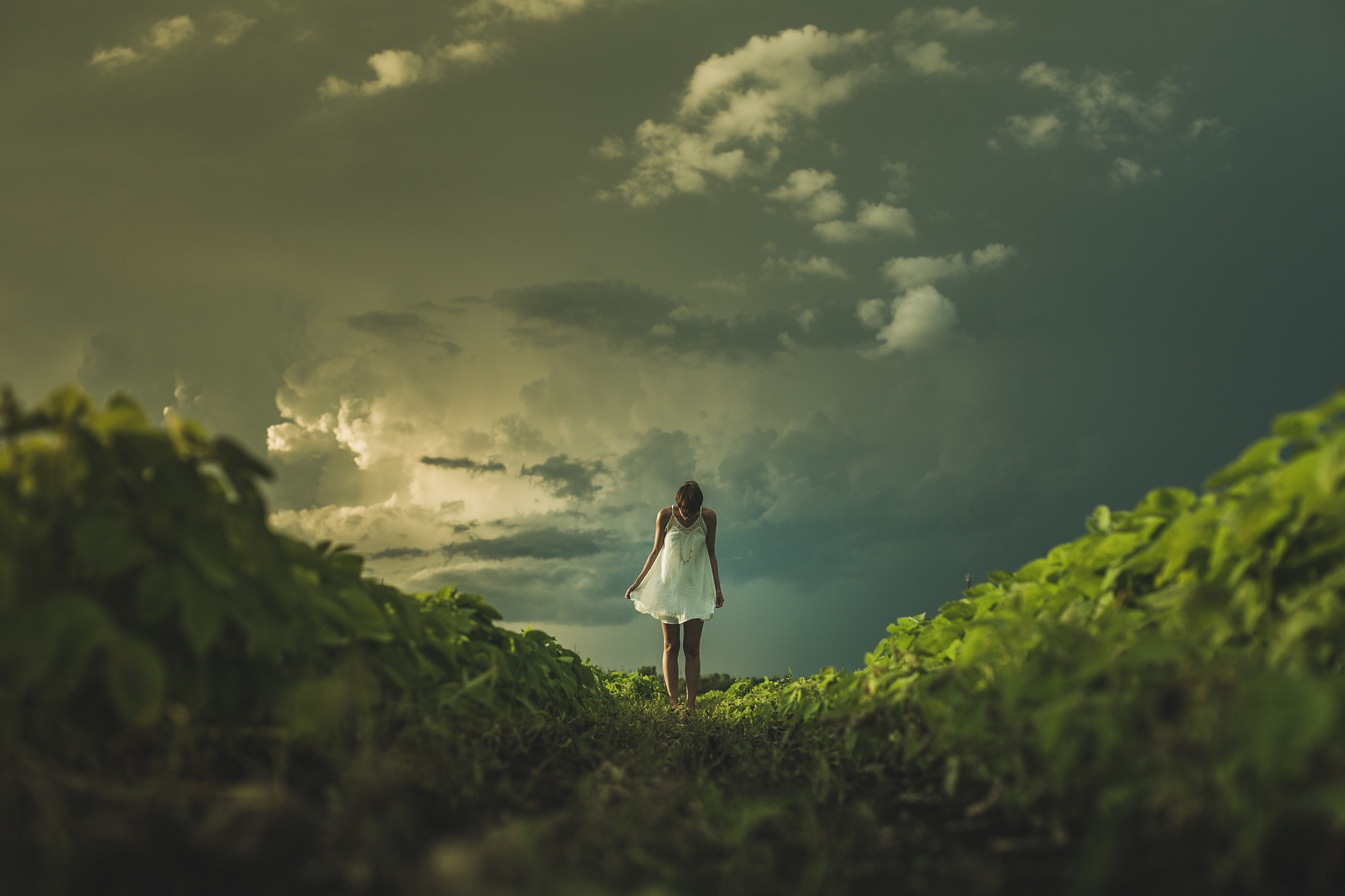 A woman in a short, white dress walking through a field under a cloudy sky.