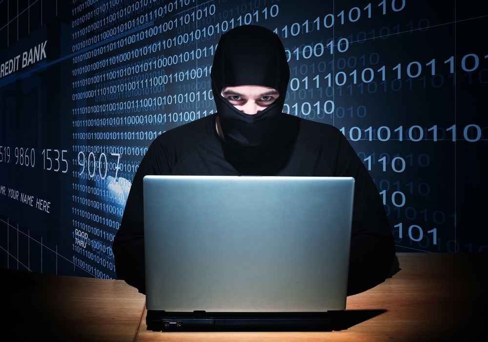A man in a ski mask working on a laptop and looking menacing. There is binary code behind him.