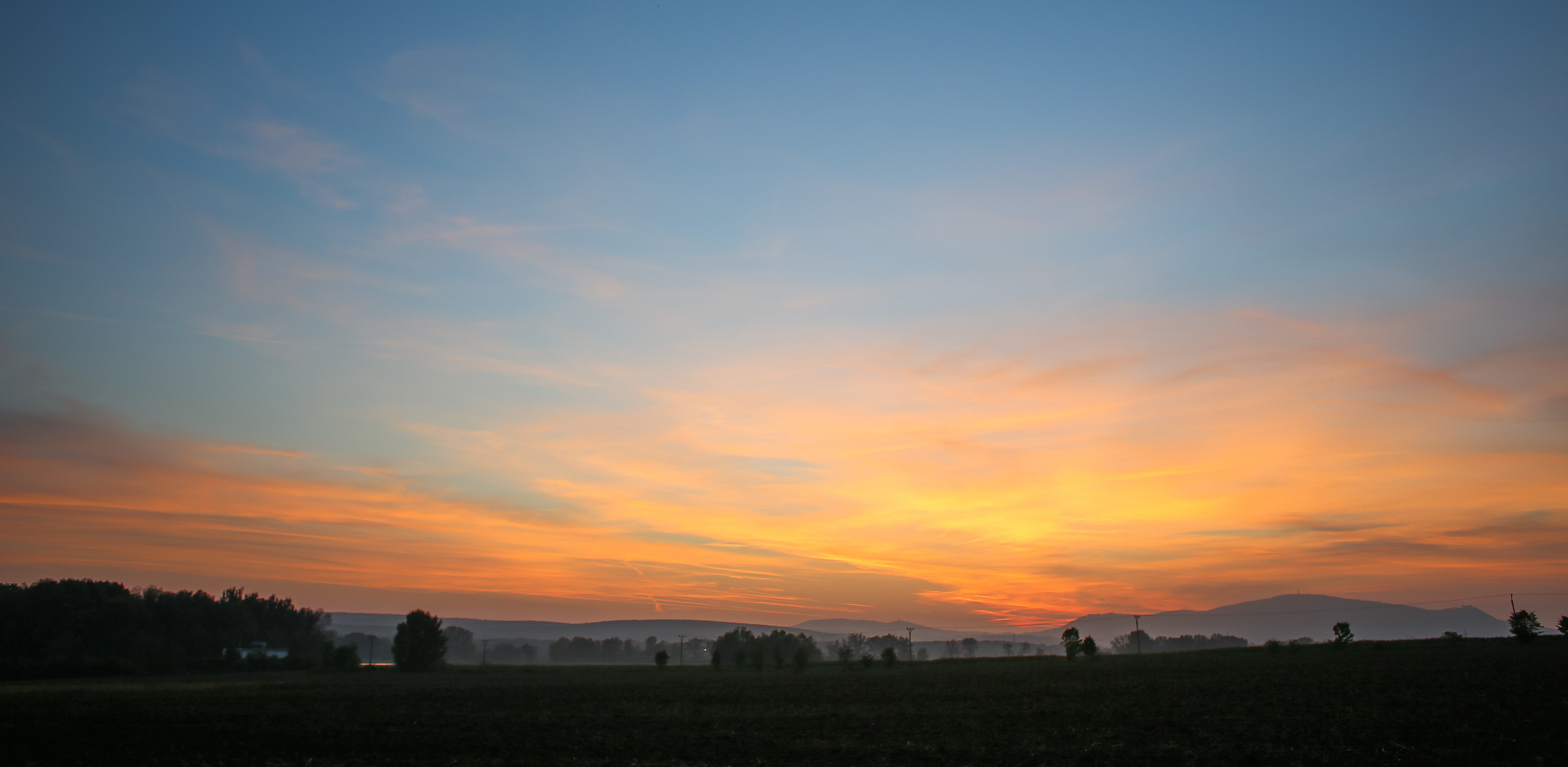 A sunset over a field with some trees and a mountain range along the horizon.