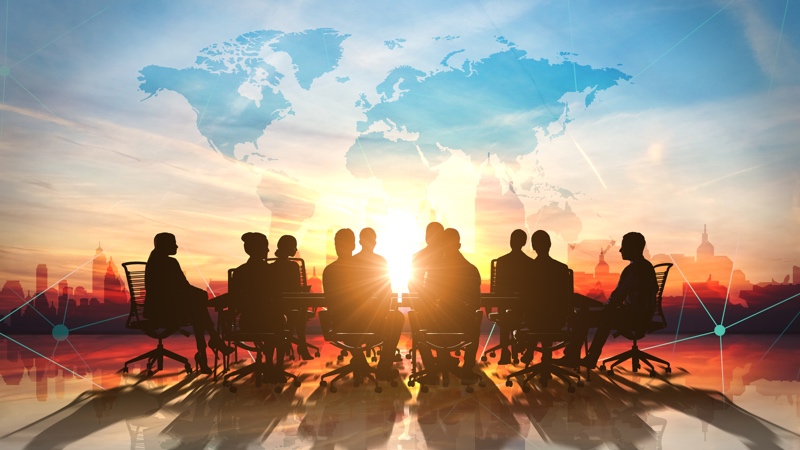 The silhouette of people sitting around a conference table with the sun coming up behind them and a city skyline.
