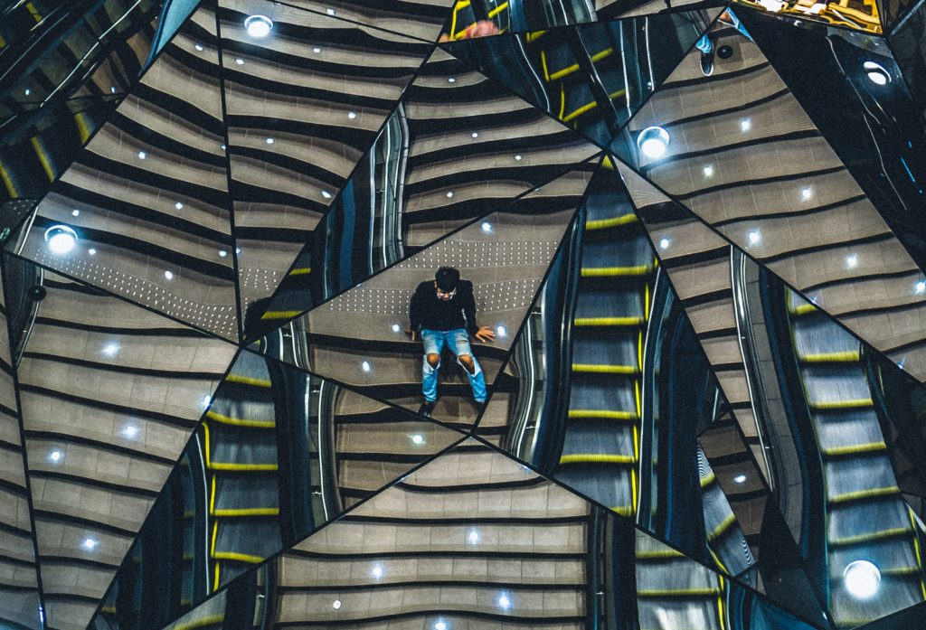 A man sitting on stairs, reflected in many triangular panels of glass.