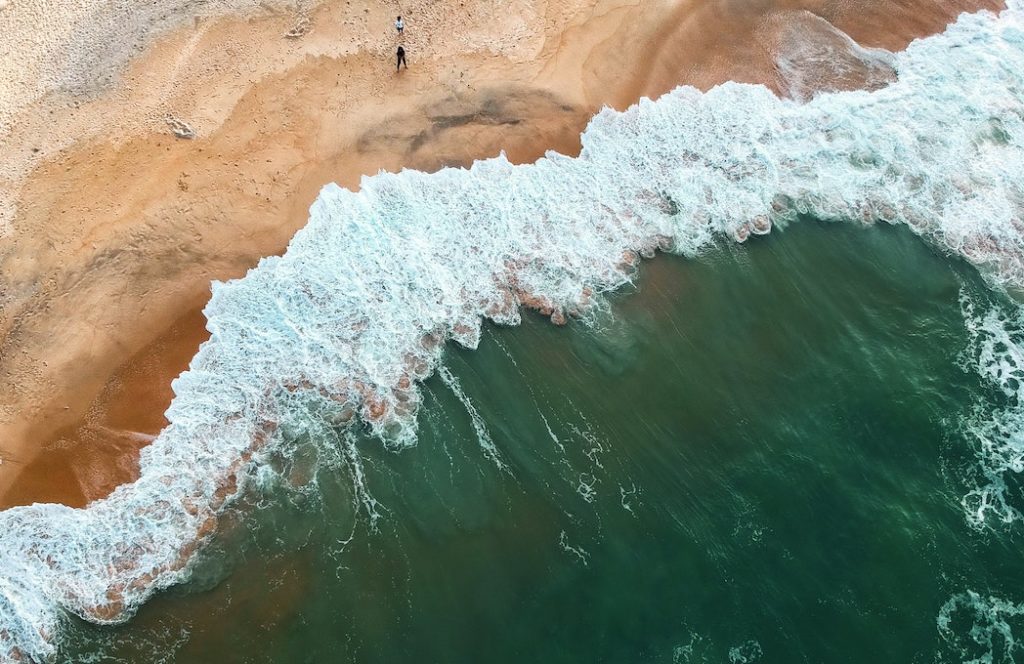 Seen from above, 2 people standing on a beach as waves roll in.