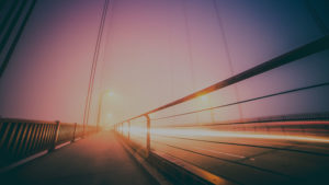 A sidewalk next to a highway on a bridge at night in the fog.