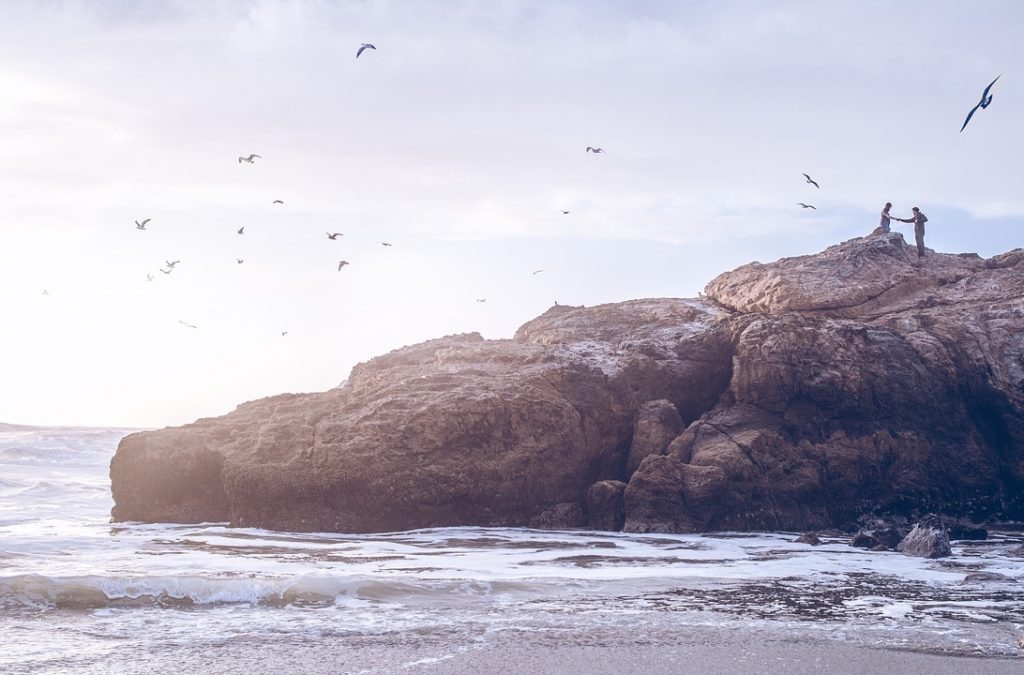 2 people standing on an outcropping of rock at the ocean with seagulls flying overhead.