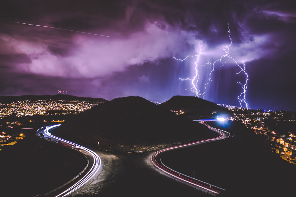 Lightning striking the earth in several places, storm clouds, cars speeding on a highway that curves around hills.