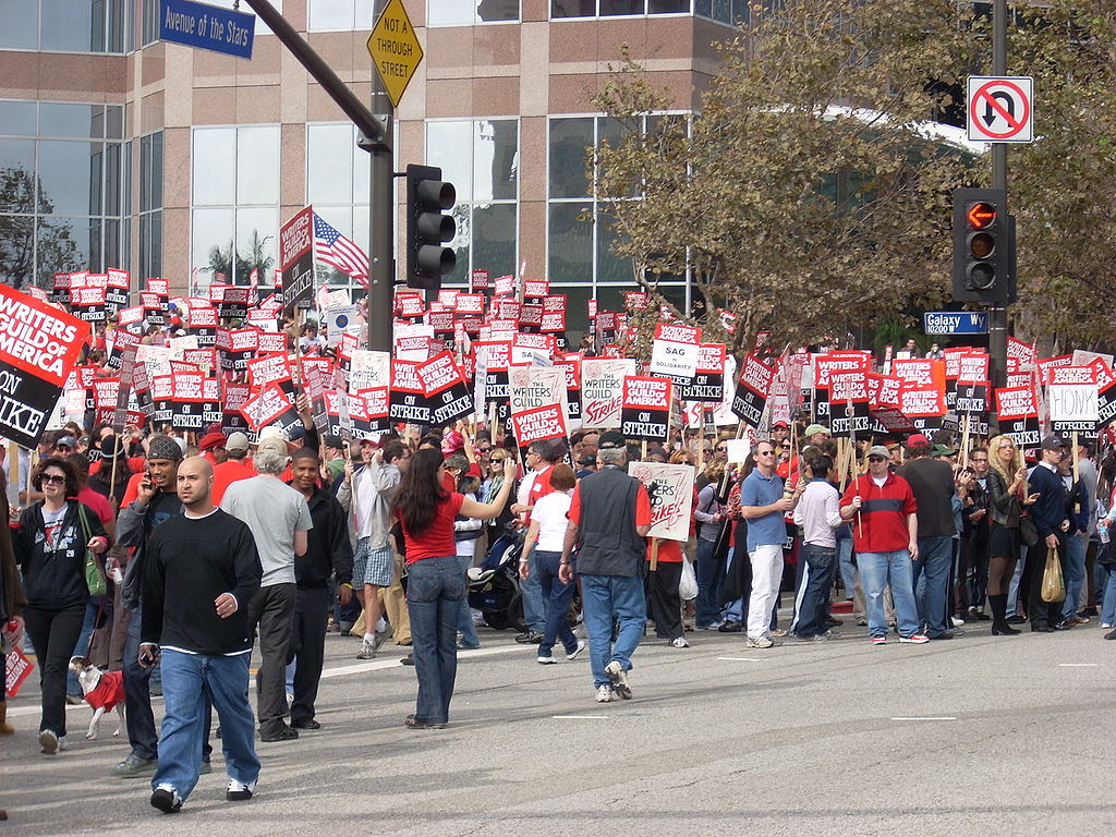 A large crowd in front of a building, holding signs that say writer's guild of America on Strike.