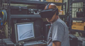 A man wearing a VR headset while standing in front of a computer with metal shelves around the room.