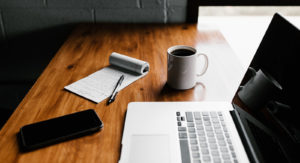 A small table with a laptop, notepad, pen, smartphone and mug of coffee.