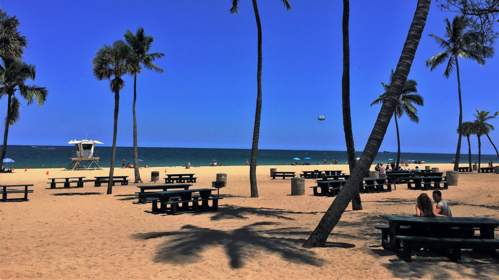 The beach in Fort Lauderdale, Florida. A person is parasailing, many are sitting at picnic tables or laying on the sand.