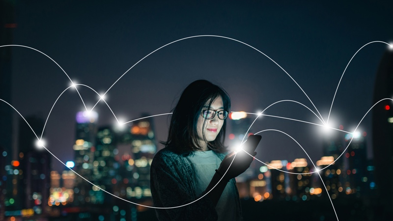 A woman looking at her phone outside at night with a city in the background and glowing, white lines all around.