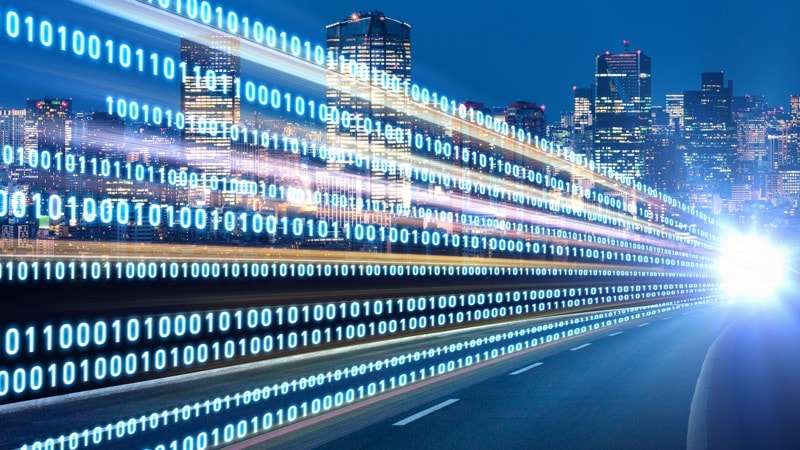 The bright lights of a car coming down a road next to a big city. There's binary code coming from the lights of the car.