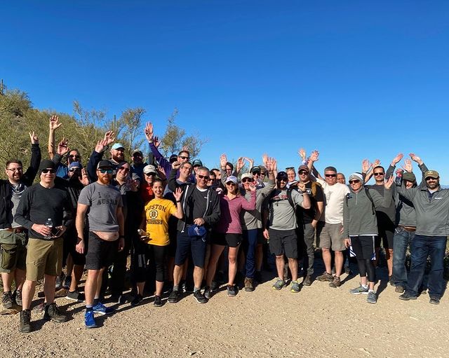 A large group of people smiling and waving at the camera. They are outside and have been hiking.