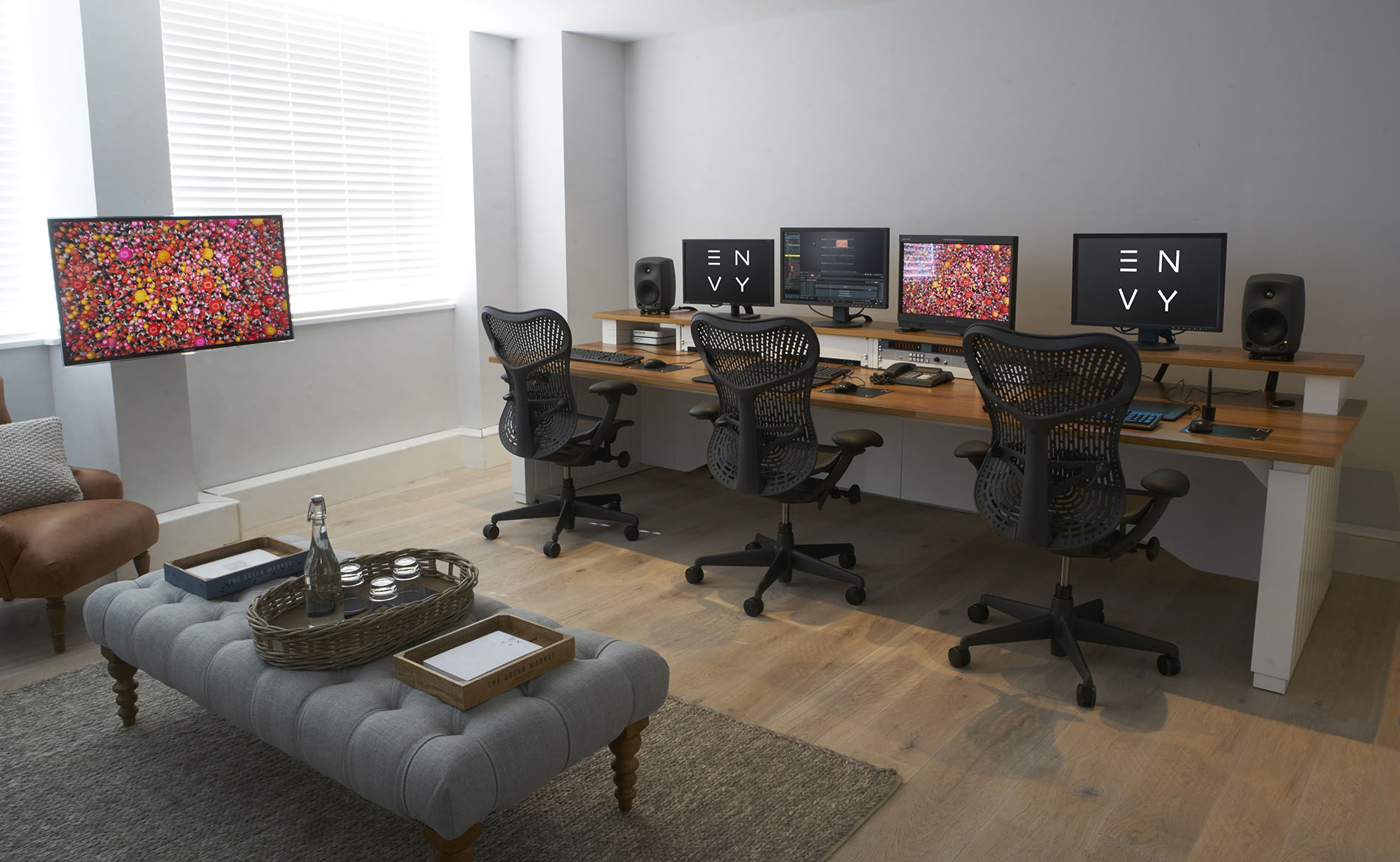 The Envy production office with a long desk with 3 chairs and computers. There's a TV monitor on the wall.