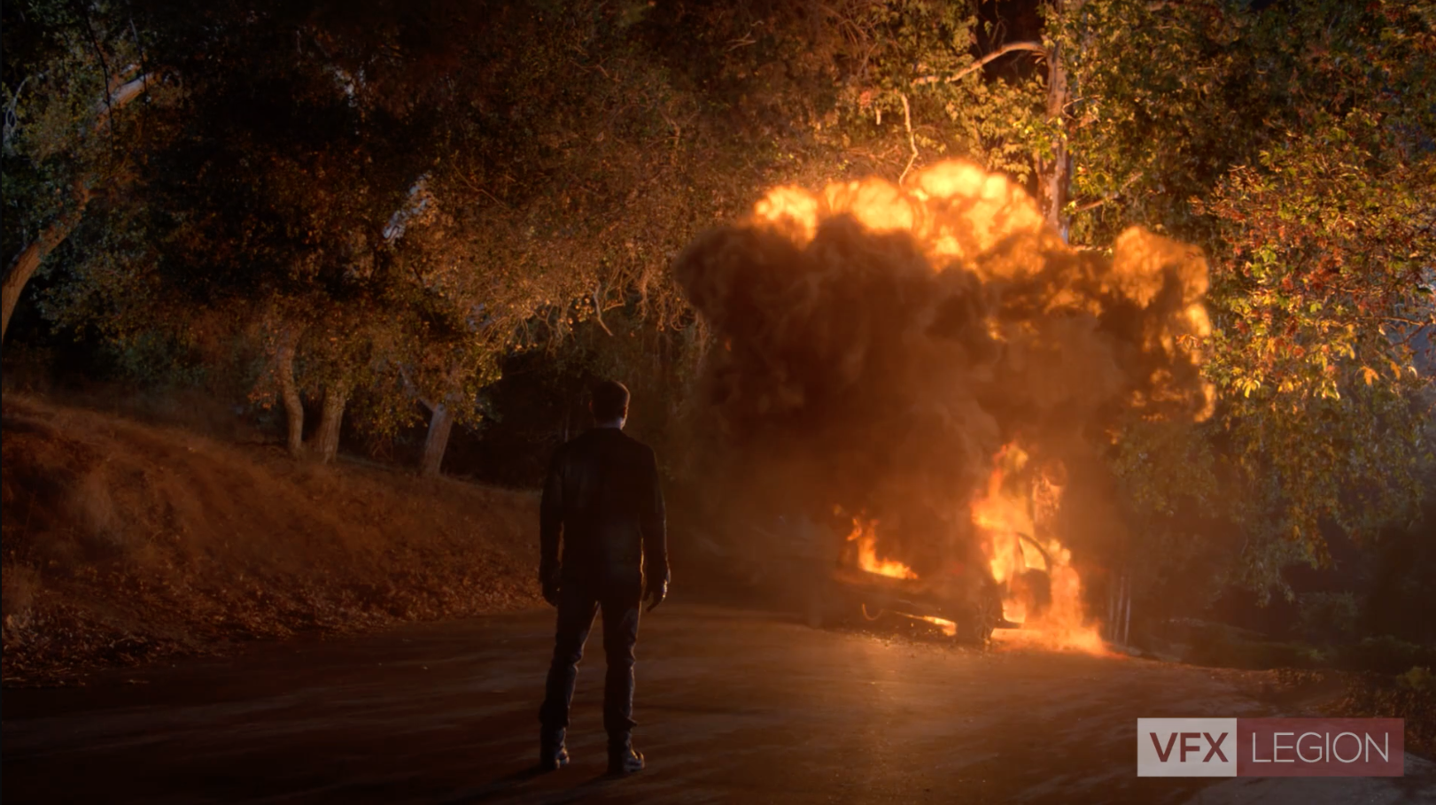 A man standing in a road, looking at a wrecked car on fire.